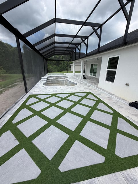 Modern patio with geometric artificial turf pattern, white pavers, screened pergola, and white house exterior with pool visible