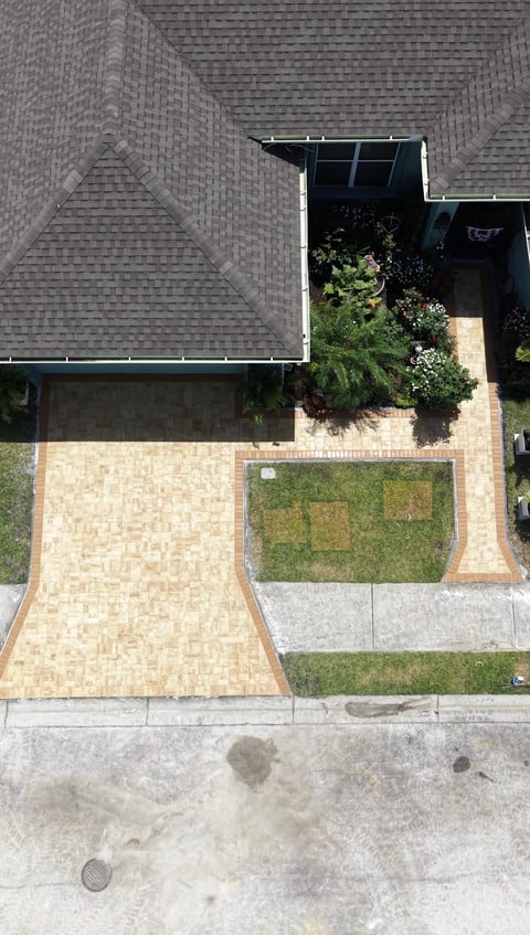 Aerial view of a residential paver driveway and front yard with grass patches and potted plants near the house entrance